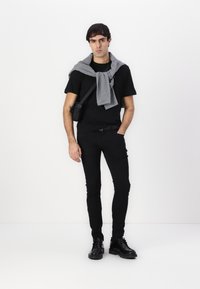 Fekete testhezálló póló, fekete slim-fit nadrág, szürke pulóver a vállakon, és fekete bakancs. Fekete válltáska az oldalon.
