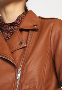 Giacca in pelle marrone con chiusura a zip e bottoni a pressione. Presenta spalline e abbinata a una maglietta nera a fantasia.