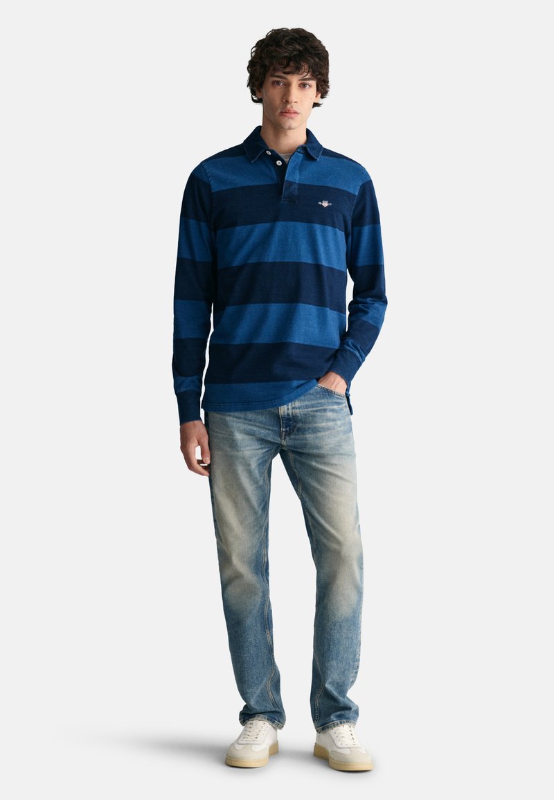 Giovane in piedi con la mano in tasca, indossa una polo a maniche lunghe a righe blu, jeans sbiaditi e sneakers bianche su uno sfondo semplice.