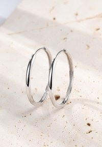 Pendientes de aro de plata con un acabado pulido. Son circulares, con un diseño suave y elegante. Presentados sobre un fondo de piedra beige texturizada.