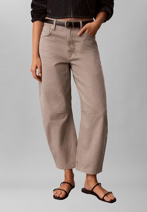 Žena oblečená v béžových džínsach s vysokým pásom a širokými nohavicami, čiernym opaskom, krátkou tmavou bundou a čiernymi sandálmi na remienky, ruka v vrecku.