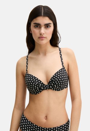 Młoda kobieta ubrana w czarny biały wzorzysty top bikini na cienkich ramiączkach, stojąca na tle gładkiego jasnoszarego tła.
