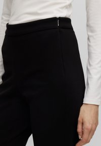 Zwarte hooggetailleerde broek met een gladde textuur, met een zijrits en een getailleerde pasvorm. Witte langemouwen top gedeeltelijk zichtbaar.