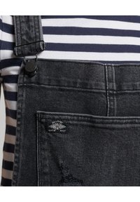 Donkerdenim tuinbroeken met verstelbare zwarte clips, versleten zakken en een merklabel. Gecombineerd met een marineblauw en wit gestreept shirt.