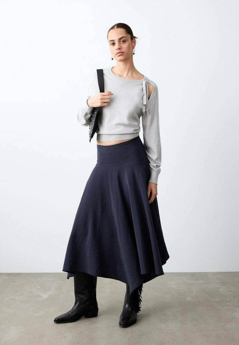 Frau trägt einen hellgrauen Cold-Shoulder-Pullover, einen asymmetrischen dunkelblauen Rock, schwarze Stiefel und hält eine schwarze, mit Nieten besetzte Handtasche über der Schulter.