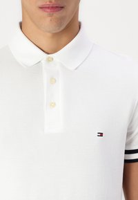 Hvit bomullspolo skjorte med en tre-knappslukking, korte ermer med svart og hvitt stripe, og en liten logo på brystet.