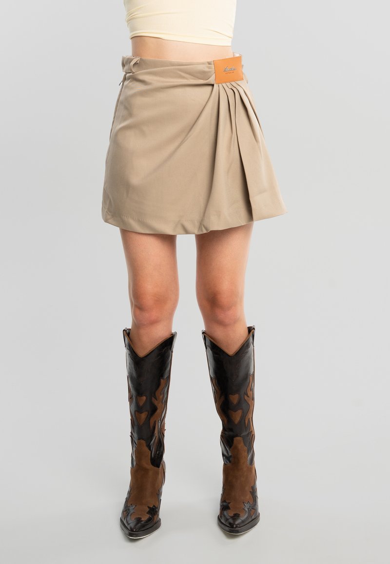 Femme portant une mini-jupe plissée beige avec une étiquette et des bottes de style western noires et marron, debout devant un fond gris.