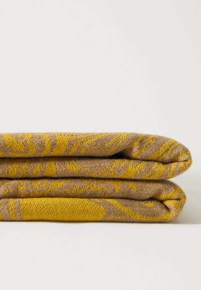 Couverture pliée jaune moutarde et beige avec un motif floral tissé complexe sur fond blanc uni.