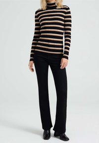 Mujer con una blusa de rayas en negro, beige y blanco de manga larga, pantalones acampanados negros y botas negras de tobillo, de pie sobre un fondo gris.