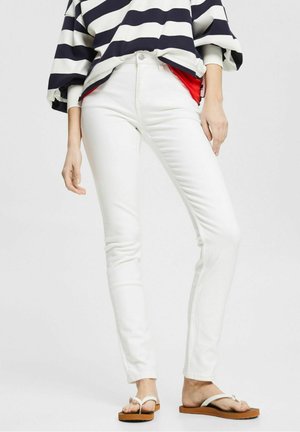 Persona con pantalones vaqueros ajustados blancos, camiseta a rayas azul marino y blanca, capa roja por debajo y chanclas blancas, de pie sobre un fondo liso.