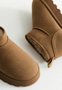 Botas de tobillo de ante marrón con punta redondeada, forro interior suave y suela de goma texturizada. Incluye detalles como una lengüeta en la parte trasera.