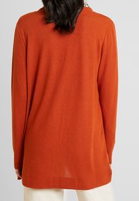 Kvinna i en långärmad, bränd orange tröja över vita byxor, stående med ryggen mot kameran mot en enhetlig bakgrund.