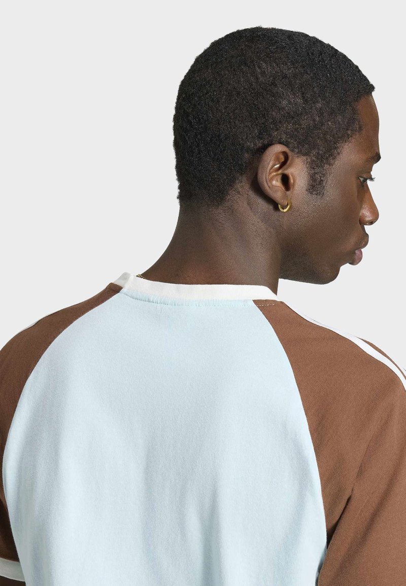 Junger Mann mit kurzen lockigen Haaren und goldenem Creolen-Ohrring, trägt ein hellblau-braunes Raglanhemd, blickt nach rechts vor schlichtem Hintergrund.