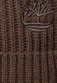 Brązowa, dziana czapka z texturowanym wzorem ściągacza. Zawiera subtelne, haftowane logo drzewa w ciemniejszym odcieniu. Miękki, ciepły materiał.