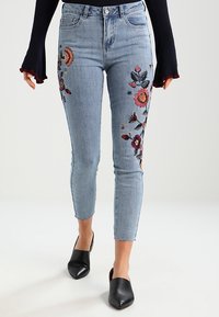 Jeans skinny de tiro alto en azul claro con bordados florales a los lados. Cuenta con un cierre de botón y dobladillos ligeramente deshilachados.