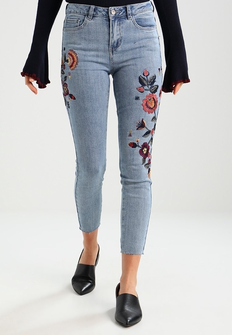 Jeans skinny de tiro alto en azul claro con bordados florales a los lados. Cuenta con un cierre de botón y dobladillos ligeramente deshilachados.
