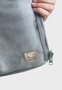 Sivi flis torba s patentnim zatvaračem, koja sadrži pravokutnu smeđu oznaku s natpisom "T-KIT" i srebrni patentni zatvarač s brend detaljima.