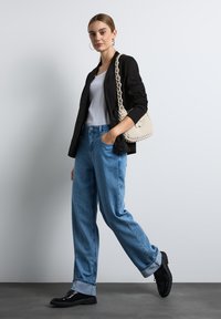 Czarna marynarka, biała bluzka na ramiączkach, jasnoniebieskie szerokie dżinsy i czarne buty. Modelka trzyma torebkę w kolorze kremowym o fakturowanym wykończeniu, pozując na tle w neutralnych odcieniach.