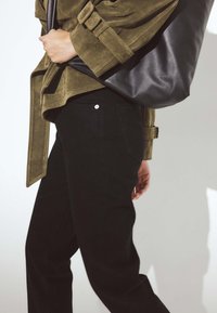 Chaqueta de ante oliva, jeans negros con un botón plateado y un gran bolso de cuero negro. Se destacan las texturas suaves y los colores neutros.