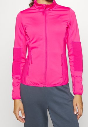 Kvinde iført en lys pink jakke med fuld lynlås, ribstrikkede albuelapper og lynlåslommer i siderne, kombineret med mørkegrå bukser.