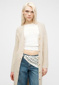 Νέα γυναίκα με μακριά ξανθά μαλλιά, φορώντας μπεζ ζακέτα με κουμπιά, λευκό αμάνικο τοπ, μπλε τζιν και λευκή ζώνη με πλεκτή λεπτομέρεια.