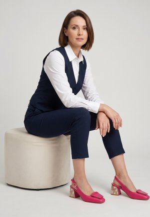 Vrouw met kort bruin haar, gekleed in een marineblauw vest en broek, wit overhemd en roze schoenen met blokhak, zittend op een ronde beige poef tegen een witte achtergrond.