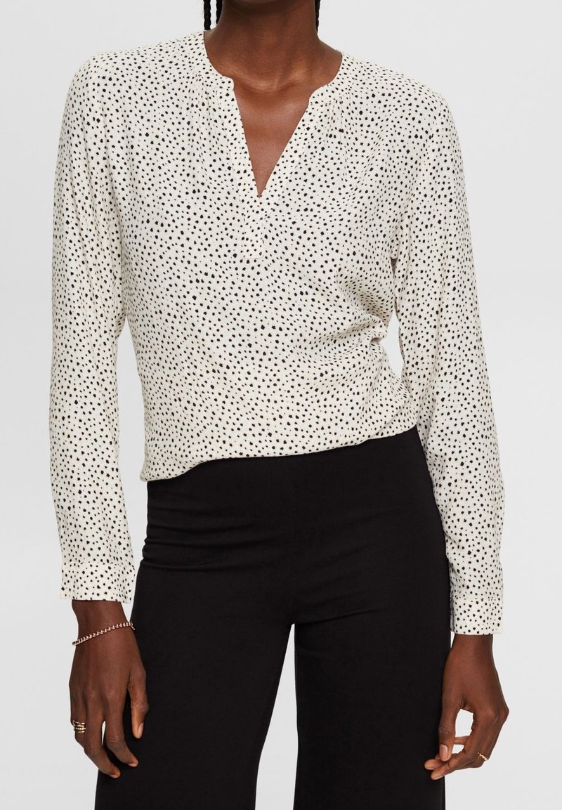 Femme portant un chemisier blanc à manches longues avec des taches noires et un pantalon noir, accessoirisée avec des bagues et un bracelet.