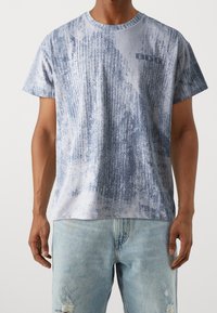 Krátký rukáv tričko v světle modré s texturovaným designem. Obsahuje malé logo na hrudi a kulatý výstřih. Materiál vypadá měkký.