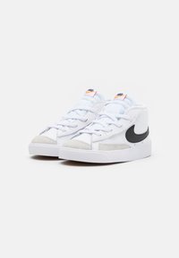 Bílé tenisky s hladkým koženým svrškem, semišovými detaily, černým logem Nike swoosh, plochou bílou podrážkou a světle modrými tkaničkami.