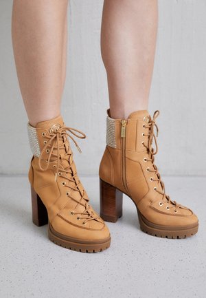 Bottes chevilles en cuir camel à lacets, avec fermeture éclair latérale et talons larges en bois, portées sur jambes nues sur un fond clair.