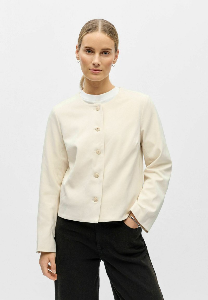 Femme aux cheveux blonds attachés en queue de cheval basse, portant une veste crème boutonnée sur un haut blanc et un pantalon noir, debout avec une main dans la poche.