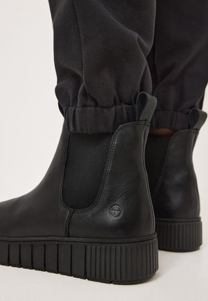 Bottines en cuir noir avec des panneaux élastiques sur les côtés, semelles en caoutchouc texturé et un logo discret. Portées avec un pantalon noir retroussé.