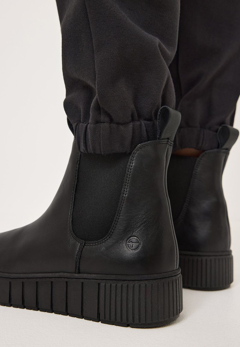 Bottines en cuir noir avec des panneaux élastiques sur les côtés, semelles en caoutchouc texturé et un logo discret. Portées avec un pantalon noir retroussé.