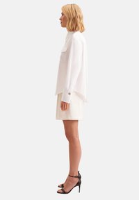 Blusa bianca con maniche lunghe, polsini con bottoni e colletto a punta, abbinata a una gonna chiara e sandali neri con tacco alto.
