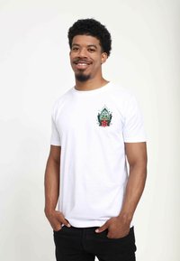 Hvid bomulds T-shirt med et livligt grønt og blomstret broderet patch på venstre bryst. Casual fit med korte ærmer. Sorte jeans.