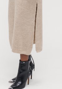 Μπεζ πλεκτό πουλόβερ με πλευρικές σχισμές, συνδυασμένο με μαύρες δερμάτινες μποτίνες μέχρι τον αστράγαλο με μυτερή μύτη, ασημένια κάπας και λεπτομέρειες από κορδόνια.