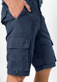 Pantaloni cargo blu navy con molte tasche, bordi arrotolati e texture liscia. Presentano cuciture visibili e dettagli con bottoni.