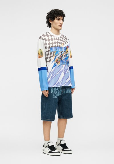 Νέος άνδρας που φοράει μακρυμάνικο μπλουζάκι με γραφικά σε μπλε και μπεζ αποχρώσεις, τζιν σορτς, λευκές κάλτσες και ασπρόμαυρα αθλητικά παπούτσια, στέκεται πλαγίως.