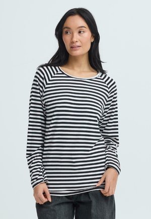 Vrouw met lang donker haar, gekleed in een zwart-wit horizontaal gestreept shirt met lange mouwen en donkere broek, kijkt naar de zijkant.