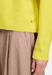 Jasnożółty, dziergany sweter z prążkowanymi mankietami i ściągaczem u dołu. Posiada mały złoty akcent w pobliżu dołu. Połączony z jasnobeżowymi spodniami.