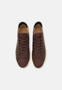 Brown leather sneakers with textured upper, rounded toe, and black heel detail. Features brown laces and a light-colored rubber sole.