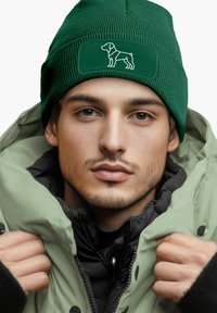 Green beanie with ribbed texture and a rectangular patch featuring a white dog silhouette. Worn with a light green puffer jacket.