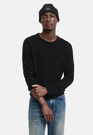 Svart langermet genser med rund hals laget av mykt materiale, sammen med falmet blå jeans. En ribbestrikket lue tilfører tekstur og varme.