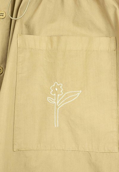 Tissu beige avec une poche carrée ornée d'une simple fleur blanche brodée avec une feuille et deux boutons sur le côté.