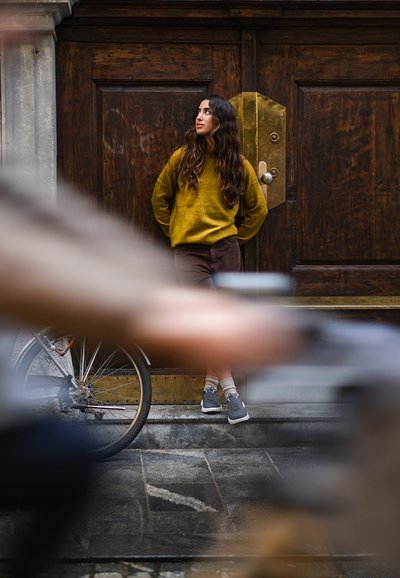 Žena se opírá o dřevěné dveře, má na sobě hořčicový svetr a hnědé kalhoty, se šedými botami a ponožkami, vedle jí stojí kolo.