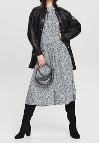 Giubbotto di pelle nera sopra un vestito midi azzurro chiaro a fantasia con vita arricciata. Tiene una borsa in grigio scuro texturizzato. Stivali neri al ginocchio.