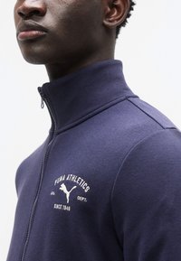 Σπορ μπουφάν σε σκούρο μπλε με φερμουάρ, ψηλό γιακά, απαλό ύφασμα, κεντητό λευκό λογότυπο "PUMA ATHLETICS" και εφαρμοστή γραμμή.