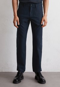 Marineblauwe taps toelopende broek van een gladde stof, met een knoopsluiting, frontzakken en een strakke, minimalistische uitstraling. Gecombineerd met zwarte veterschoenen.
