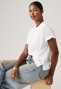 Λευκό βαμβακερό μπλουζάκι με ρεβέρ στα μανίκια, με άνετη εφαρμογή. Συνδυασμένο με ανοιχτό μπλε, ξεθωριασμένο τζιν φαρδιάς γραμμής, τοποθετημένο πάνω σε ξύλινη επιφάνεια.