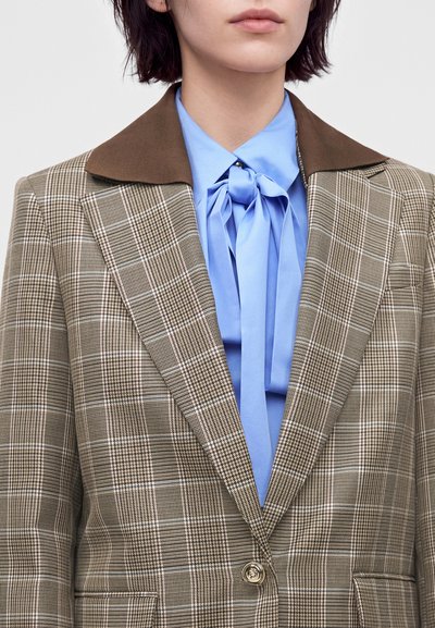 Femme portant un blazer à carreaux marron avec un large col sur un chemisier bleu clair noué en nœud au cou, cadrée à la bouche et à la poitrine.